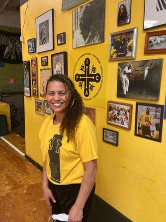 photo of Contra Mestra Andrea in front of a wall of photos of capoeira practitioners